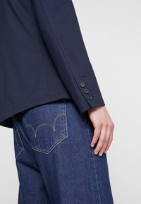 Námornická blazer s hladkým povrchem, s jedním řadou tří knoflíků na manžetě, spárovaný s tmavými džínami se žlutými švy.