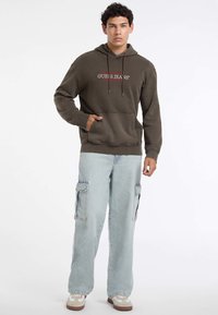 Giovane uomo in piedi con una felpa marrone di Guess, jeans cargo azzurri chiari e sneakers bianche su uno sfondo bianco.