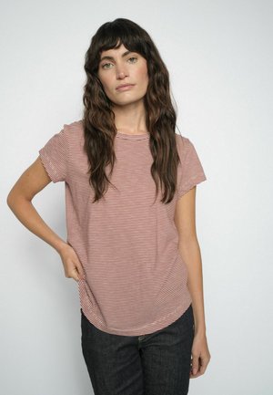 Vrouw met lang golvend haar, gekleed in een rood-wit gestreept shirt met korte mouwen en een donkere broek, staand tegen een effen lichte achtergrond.