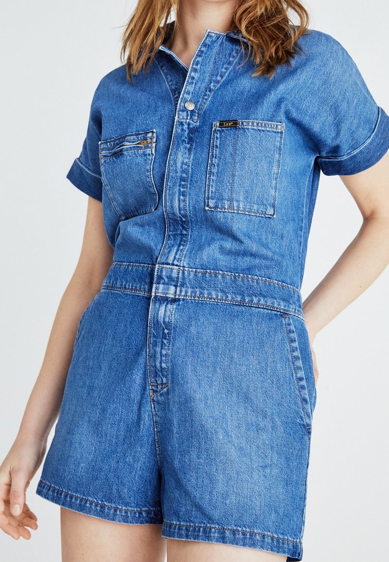 Femme portant une combinaison short en denim bleu à manches courtes avec fermeture à boutons sur le devant et poches poitrine, debout avec une main sur la hanche.