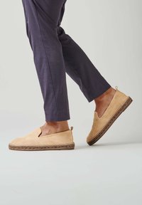 Chaussures slip-on en daim tan avec une semelle en jute tissée et des découpes sur les côtés, assorties à un pantalon tapered bleu marine, présentant un design décontracté.