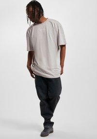 Light gray short-sleeve t-shirt with a round neckline and embroidered logo on the chest, paired with dark denim jeans and grey sneakers.
