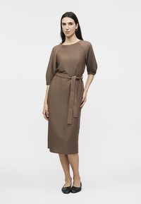 Femme portant une robe midi côtelée marron avec des manches trois-quarts et une ceinture, debout devant un fond clair uni.