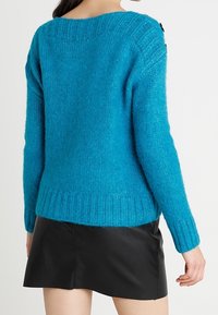 Maglione turchese in maglia pelosa con orlo a coste e maniche testurizzate, caratterizzato da bottoni decorativi sulla spalla. Indossato con una gonna di pelle nera.