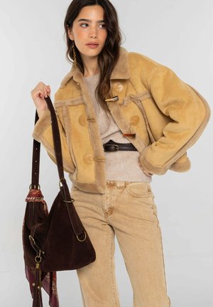 Femme portant une veste en peau retournée beige, un pull beige, un jean marron clair, tenant un sac en daim marron foncé avec un foulard à motifs attaché à la sangle.