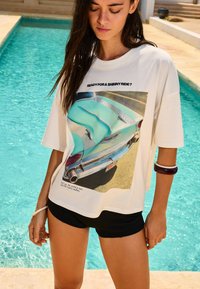 T-shirt graphique blanc arborant un design de voiture rétro imprimé et le texte "PRÊT POUR UN RIDE ÉBLOUISSANT ?" Associé à un short noir.