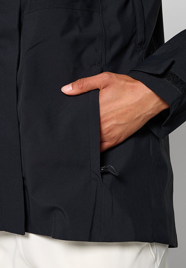 Ręka częściowo wsunięta w czarną kieszeń kurtki z regulowanym paskiem na nadgarstek, noszonej na białych spodniach.