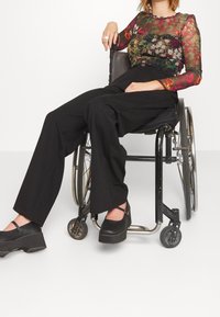 Top fleuri transparent à manches longues en plusieurs couleurs, associé à un pantalon noir taille haute et des chaussures à plateforme noires. Assise dans un fauteuil roulant.