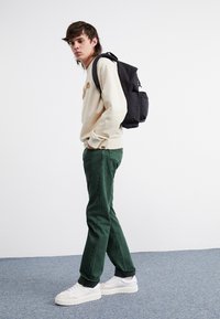 Beige mikina s logem, zelené džíny a bílé tenisky. Černý batoh přes jedno rameno, umístěný na jednobarevném bílém pozadí.