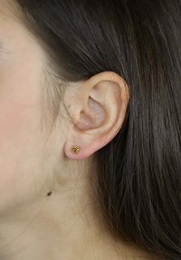 Złoty kwiatowy kolczyk na płatku ucha, z detalicznym wzorem kwiatu. Włosy proste i brązowe, częściowo zakrywające ucho.