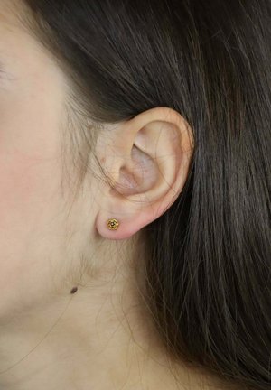 Złoty kwiatowy kolczyk na płatku ucha, z detalicznym wzorem kwiatu. Włosy proste i brązowe, częściowo zakrywające ucho.