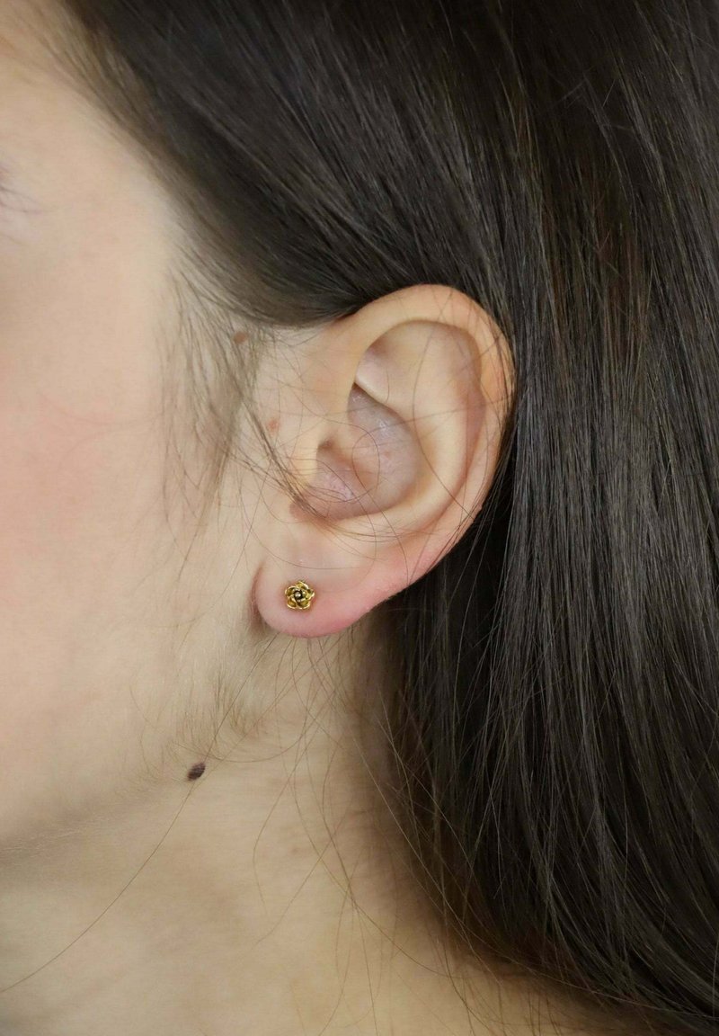 Złoty kwiatowy kolczyk na płatku ucha, z detalicznym wzorem kwiatu. Włosy proste i brązowe, częściowo zakrywające ucho.