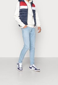 Vadderad marinblå väst med röda och vita ränder, vit hoodie, ljusblå jeans och marinblå skor med vita inslag; avslappnad outfit.