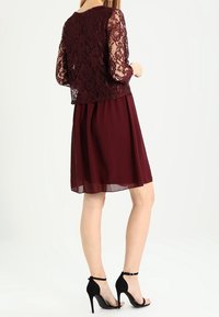 Burgundiklänning med spetsöverdel och långa ärmar, som har en flödande chiffonskjol. Svarta stilettklackar kompletterar outfiten.