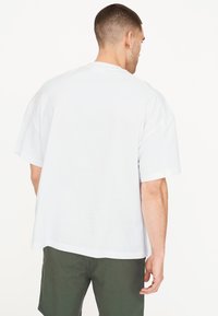 Camiseta de algodón blanca de manga corta, con un corte relajado y cuello redondo. Se lleva con pantalones cortos verdes oscuros, mostrado desde la parte de atrás.