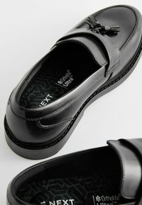 Chaussures noires en cuir avec une semelle plate, présentant une large bande sur le dessus et un gland décoratif. Intérieur Ortholite® Ultra.
