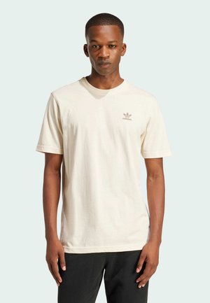 T-shirt di cotone crema con maniche corte, colletto rotondo e un piccolo logo marrone adidas sul petto. Indossato con pantaloni neri.