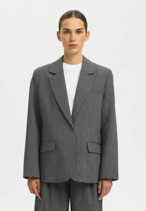 Femme aux cheveux tirés en arrière portant un large blazer gris à fines rayures avec un pantalon assorti, sur un haut blanc, debout devant un fond uni.