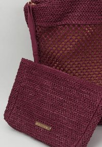 Bolso tejido burdeos con un diseño de malla transparente, que presenta una textura suave y un estuche a juego desmontable con una etiqueta de logo.