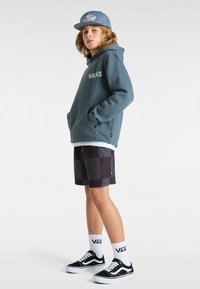 Sudadera con capucha en color azul verdoso con el logo "VANS", combinada con pantalones cortos a cuadros negros, calcetines blancos y zapatillas de skate negras. Complementada con una gorra gris.