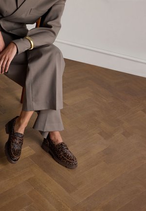 Brown leopard print loafers with a chunky sole, paired with tailored gray trousers. Wooden herringbone floor in the background.