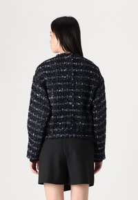 Marineblauwe tweed jasje met textuur, losse pasvorm, brede mouwen en een verkorte lengte, gecombineerd met een zwarte minirok.