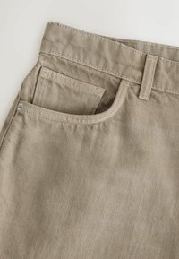 Pantalon en denim beige en gros plan montrant une poche avant avec une petite poche à monnaie à l’intérieur et une passants de ceinture au-dessus sur un fond clair.