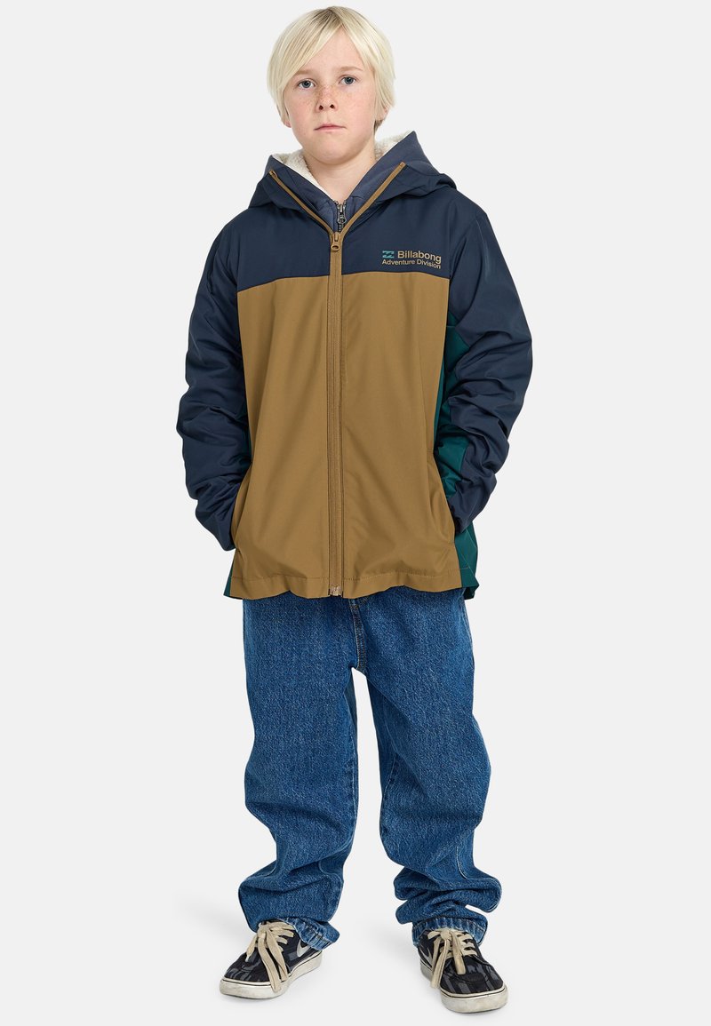 Giacca con cappuccio marrone, blu e verde, con zip frontale, polsini elasticizzati e logo Billabong. Indossata su jeans blu larghi e sneakers.