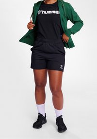 Camiseta Hummel negra con logo blanco, chaqueta verde con cremallera, pantalones cortos negros y zapatillas negras con acentos blancos. El modelo lleva calcetines blancos.