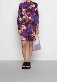 Lila Kleid mit floralem Muster, langen Ärmeln und verstellbarem Taillengürtel, kombiniert mit schwarzen Plateauslippern und einer kleinen lavendelfarbenen Handtasche.