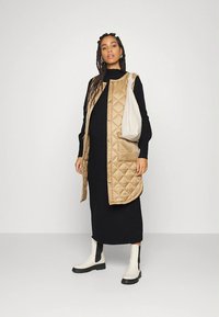 Beżowa pikowana kamizelka z przodu na guziki, noszona na czarnej prążkowanej sukience. Białe botki z czarnymi akcentami. Modelka trzyma jasną torebkę.