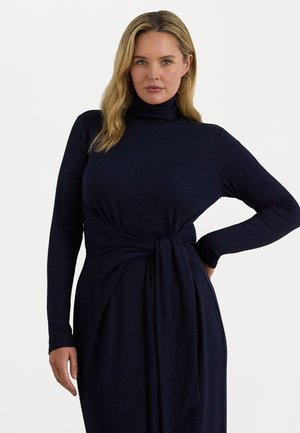 Vrouw met blond haar die een donkerblauwe lange mouw turtleneck jurk draagt met een gestrikte tailleband, poseert tegen een effen witte achtergrond.