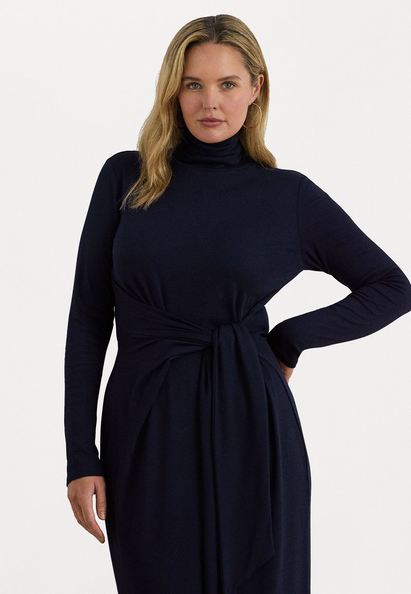 Donna con capelli biondi che indossa un vestito a maniche lunghe blu scuro con colletto alto e cintura annodata in vita, in posa contro uno sfondo bianco uniforme.