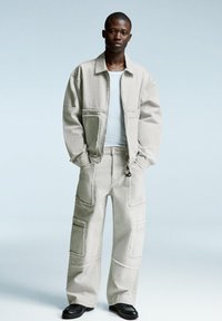 Young man standing with hands in pockets wearing a beige oversized jacket and matching patch-pocket pants, with black shoes.