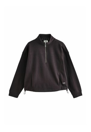 Femme blonde aux cheveux bouclés portant un pull noir zippé avec le texte "self." et des manches longues rouges, debout en plein soleil.