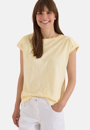 Jeune femme souriante, vêtue d'un haut jaune clair à manches courtes et d’un pantalon blanc, debout avec une main dans la poche sur un fond neutre.