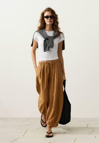 Femme portant des lunettes de soleil, un t-shirt blanc, un pantalon marron ample, un pull rayé drapé sur les épaules, portant un sac noir, marchant sur un sol carrelé.