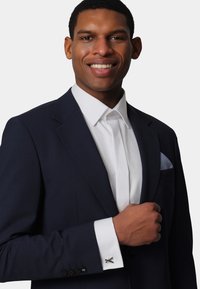 Navy suit with notched lapels, white shirt with a structured collar, and silver cufflinks. Features a light blue pocket square.