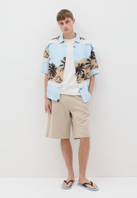 Camisa de manga corta en azul claro con estampado de palmeras y montañas, combinada con pantalones cortos beige y chanclas negras con acentos azules.