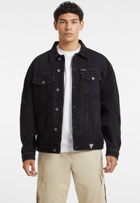 La giacca di jeans nera presenta una chiusura frontale con bottoni, due tasche sul petto e una vestibilità rilassata. Tra gli elementi distintivi ci sono bottoni argentati e una toppa con logo.