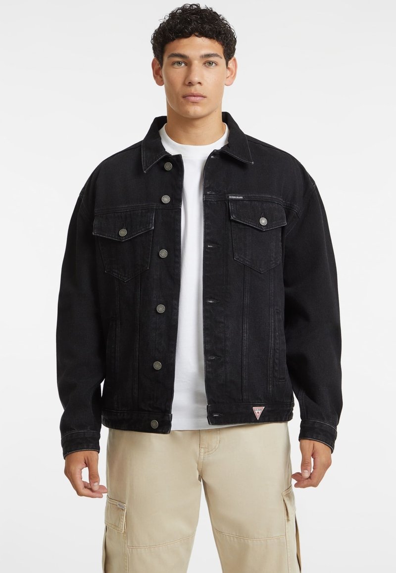 La giacca di jeans nera presenta una chiusura frontale con bottoni, due tasche sul petto e una vestibilità rilassata. Tra gli elementi distintivi ci sono bottoni argentati e una toppa con logo.