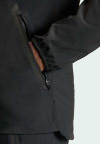 Chaqueta negra con una textura suave, diseño ajustado y detalles de bolsillos con cremallera en los laterales, con puños elásticos para un ajuste ceñido.