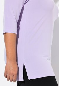 Tunika lilla con maniche 3/4, texture morbida, spacchi laterali e orlo curvo. Abbinata a pantaloni neri, che evidenziano una vestibilità rilassata.