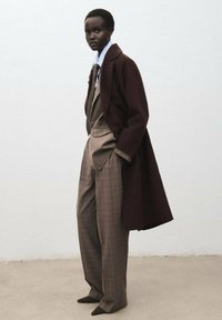 Manteau marron sur un costume à carreaux dans des tonalités beige et marron. Le costume se compose de pantalons larges et d'une chemise à col pointu. Tissus texturés.