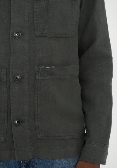 Veste à boutons couleur kaki en tissu texturé, avec poches frontales et une petite étiquette de marque. Présente des boutons noirs et une coupe décontractée.