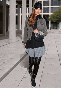 Graue Jeansjacke über einem schwarzen Strickpullover und einem schwarz-weißen karierten Hemdkleid, kombiniert mit schwarzen Lederhosen und dunklen Sneakers.