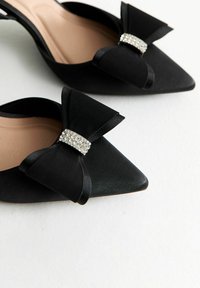 Ballerines noires à bout pointu avec de grands nœuds en satin et des embellissements en rhinestone sur fond blanc.