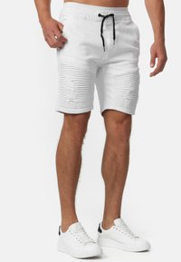 Pantalones cortos de mezclilla blanca con líneas horizontales texturizadas, acentos desgastados, una cinturilla elástica y un cordón negro. Combinados con zapatillas blancas.