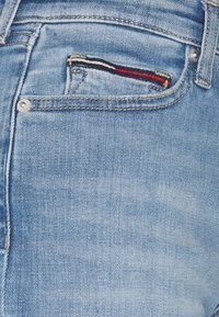 Gros plan sur un jean en denim bleu clair montrant une petite poche avant avec une étiquette en tissu présentant des rayures rouges, blanches et noires.