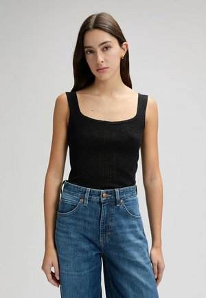 Giovane donna con capelli lunghi e lisci, che indossa una canotta nera senza maniche e jeans blu a vita alta, in piedi davanti a uno sfondo semplice.